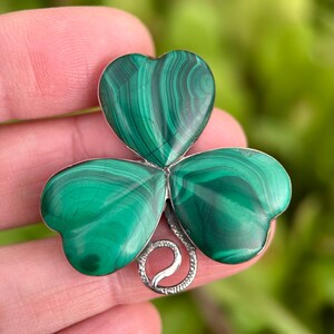 May include: A green malachite clover-shaped brooch with a silver-colored metal base. The brooch features three heart-shaped leaves with swirling patterns of dark and light green. The metal base has a decorative swirl design.