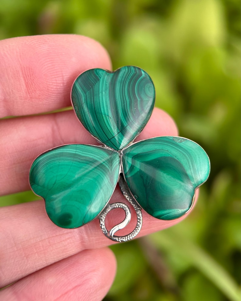 May include: A green malachite clover-shaped brooch with a silver-colored metal base. The brooch features three heart-shaped leaves with swirling patterns of dark and light green. The metal base has a decorative swirl design.