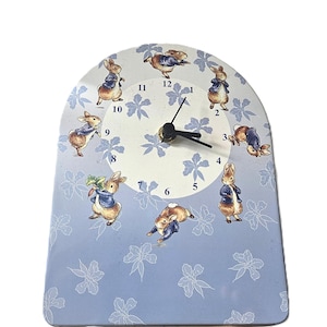 May include: A decorative clock featuring illustrations of rabbits in various poses. The clock face is light blue with white floral patterns and the numbers 1 through 12. The clock hands are black.