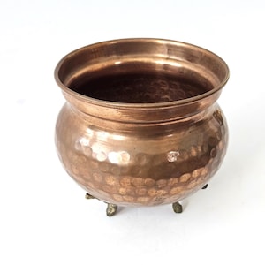 May include: A copper-colored, hammered metal pot with a rounded body and a wide, flared rim. The pot sits on three small, decorative feet. The surface has a textured, dimpled pattern. The interior is dark.