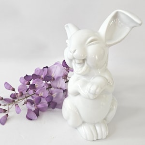 May include: A glossy white ceramic bunny figurine with a smiling expression, crossed paws, and long ears. A sprig of purple wisteria flowers is next to the bunny. The figurine is a decorative item.