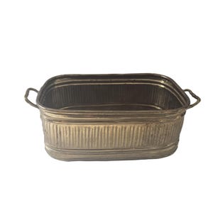 May include: A rectangular, galvanized metal tub with two curved handles. The tub has vertical ribbed detailing on the sides and a slightly rounded base. The metal has a weathered, antique appearance.