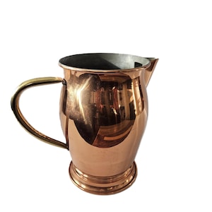 May include: A copper-coloured jug with a rounded body, a spout, and a brass-coloured handle. The jug has a shiny, reflective surface. The interior appears to be a darker metal. The jug sits on a small, circular base.