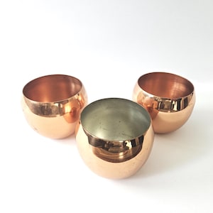 May include: Three copper-colored, spherical bowls. Two bowls have a shiny, reflective copper exterior and interior. The third bowl has a copper exterior and a silver-colored interior. The bowls are arranged on a white surface.
