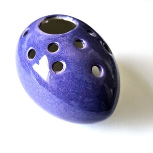 Könnte beinhalten: Eine violette, eiförmige Vase aus Keramik mit mehreren kleinen Löchern für Blumenstiele.
