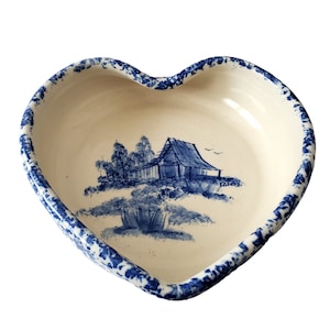 May include: Heart-shaped ceramic dish with a blue and white rural landscape design. The rim has a blue dotted pattern. The interior features a blue illustration of a house, trees, and foliage. A decorative piece for serving or display.