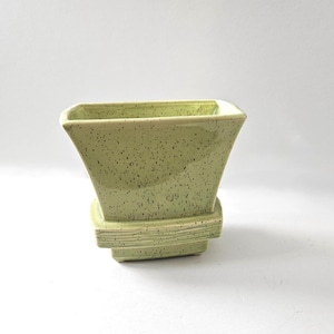 Puede incluir: Una maceta de cerámica verde claro con acabado moteado y parte superior cuadrada. La maceta tiene forma acampanada y se asienta sobre una base a juego con crestas horizontales. El diseño es simple y moderno.
