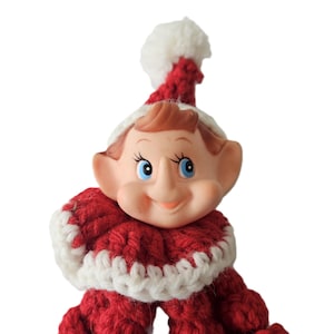 May include: A vintage Christmas elf figurine with a smiling face, blue eyes, and large ears. The elf wears a red and white crocheted hat and collar, and a red crocheted outfit. The figurine is set against a white background.