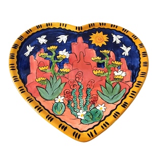 Peut inclure: Une assiette en céramique en forme de cœur avec un motif peint coloré représentant un paysage désertique. L'assiette présente des formations rocheuses rouges, des cactus verts, des fleurs blanches et un ciel bleu avec des oiseaux blancs. L'assiette a une bordure jaune avec un motif répétitif.