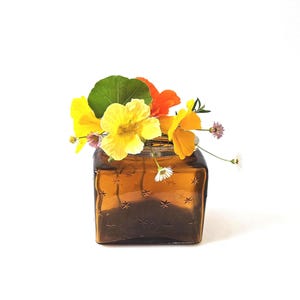 May include: A square, amber-colored glass vase filled with a vibrant arrangement of flowers. The flowers include bright yellow and orange blooms, green leaves, and small white daisies. The vase has a starburst pattern.