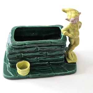 Könnte beinhalten: Ein grüner Keramik-Pflanztopf in Form eines Ziegelbrunnens mit einem kleinen gelben Eimer an der Seite. Eine gelbe Keramik-Elfenfigur lehnt sich an den Brunnen.