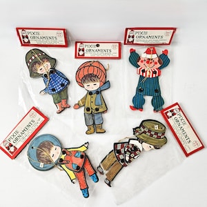 Puede incluir: Adornos Pixie vintage con ilustraciones encantadoras de niños con varios atuendos. Los adornos están empaquetados en cajas rojas y blancas, con la inscripción "Pixie Ornaments". Los niños llevan sombreros, abrigos y otros accesorios.