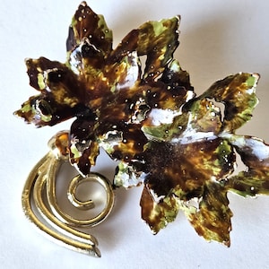 May include: A gold-tone brooch with a stylized leaf design. The leaves are brown, green, and white with a speckled finish. The brooch has a decorative scroll design at the bottom.