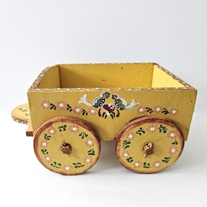 Puede incluir: Un pequeño carro de madera amarillo con detalles florales y de pájaros pintados. El carro tiene dos ruedas grandes con diseños florales rosas y verdes. El cuerpo del carro es rectangular con un borde decorativo.