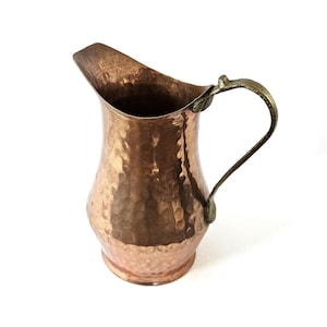 May include: A copper pitcher with a hammered texture and a brass handle. The pitcher has a wide spout and a rounded body. The handle is curved and attached to the top of the pitcher. The copper has a warm, reddish-brown color.