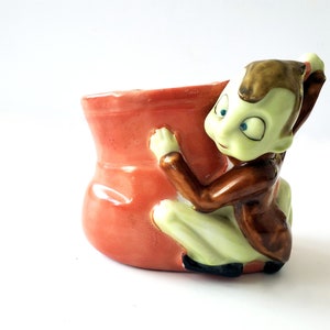 Peut inclure: Un pot en céramique en forme de sac rouge avec une figurine d'elfe verte tenant le sac. L'elfe porte une veste brune et a de grands yeux bleus.