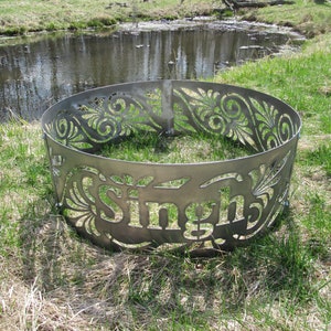 May include: A circular metal fire pit ring featuring intricate laser-cut floral patterns and the name Singh prominently displayed in the center. The gray steel structure is composed of curved panels joined by bolts, creating a decorative border.