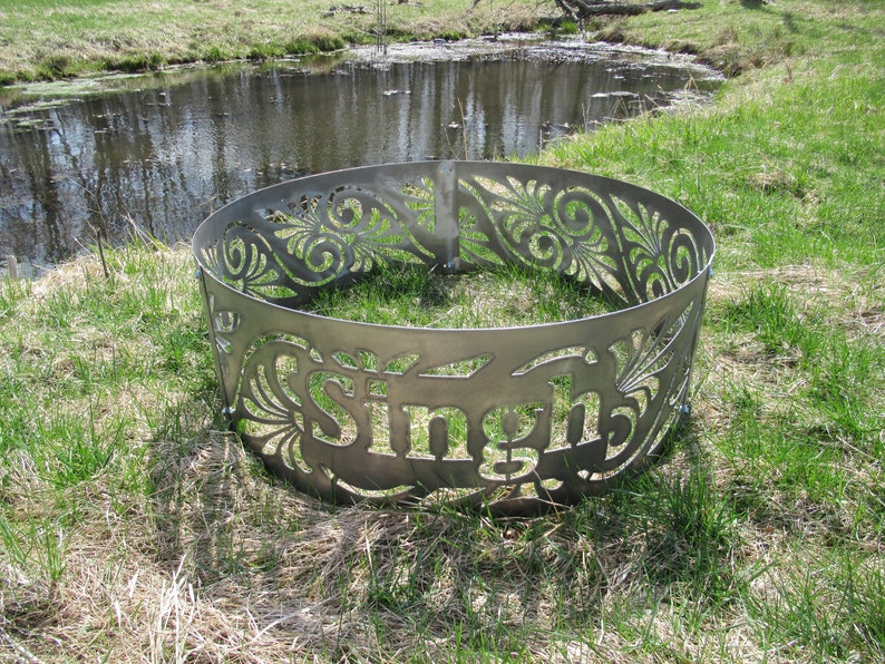 May include: A circular metal fire pit ring featuring intricate laser-cut floral patterns and the name Singh prominently displayed in the center. The gray steel structure is composed of curved panels joined by bolts, creating a decorative border.