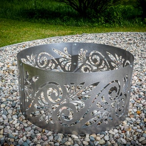 May include: A circular silver-colored metal fire pit ring featuring a laser-cut decorative pattern of swirling leaves and vines. The structure is composed of multiple curved panels connected by visible metal bolts along the vertical seams.
