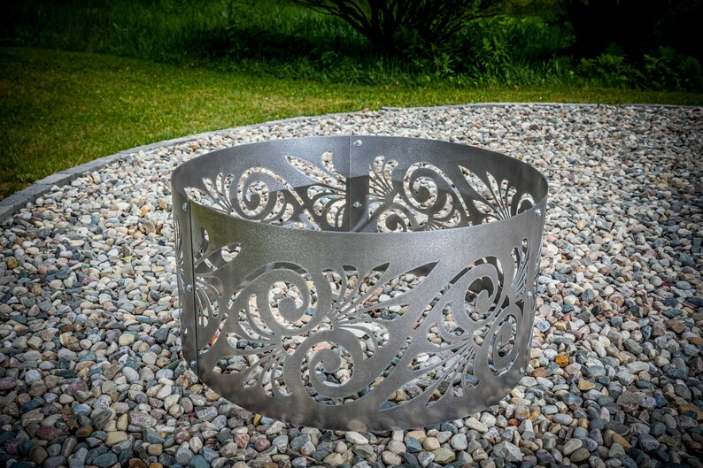 May include: A circular silver-colored metal fire pit ring featuring a laser-cut decorative pattern of swirling leaves and vines. The structure is composed of multiple curved panels connected by visible metal bolts along the vertical seams.