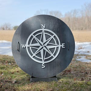 May include: A large, circular metal fire pit with a compass rose design cut out of the center. The compass rose is white, and the fire pit is dark gray. The fire pit is outdoors, in a grassy area.