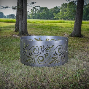 May include: A circular metal fire pit ring featuring a silver-toned finish and intricate laser-cut scrollwork patterns throughout the exterior wall. The structure is composed of multiple curved panels secured together with visible metal bolts.
