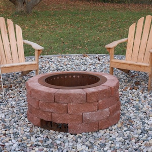 Raw steel DIY smokeless fire pit kit with no fire and visible rusty patina from outdoor exposure which is a near match to the red bricks of the fire pit with two chairs in a back yard on a gravel patio.