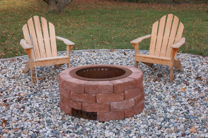 Raw steel DIY smokeless fire pit kit with no fire and visible rusty patina from outdoor exposure which is a near match to the red bricks of the fire pit with two chairs in a back yard on a gravel patio.