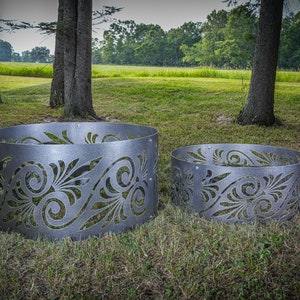 May include: Two circular metal fire pit rings featuring a laser-cut decorative pattern of swirling leaves and scrolls. The rings are constructed from grey steel panels joined by visible bolts, showcasing a repeating organic design.