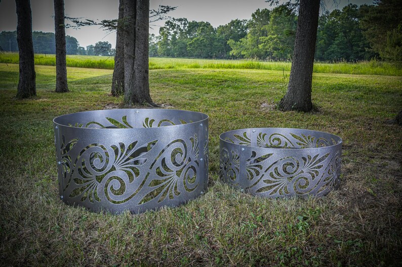 May include: Two circular metal fire pit rings featuring a laser-cut decorative pattern of swirling leaves and scrolls. The rings are constructed from grey steel panels joined by visible bolts, showcasing a repeating organic design.