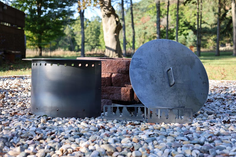 Close-up of a DIY smokeless fire ring kit with pre-cut holes and welded lip, solid steel vent blocks, and solid lid with single handle in front of stacked retaining wall blocks on gravel patio.
