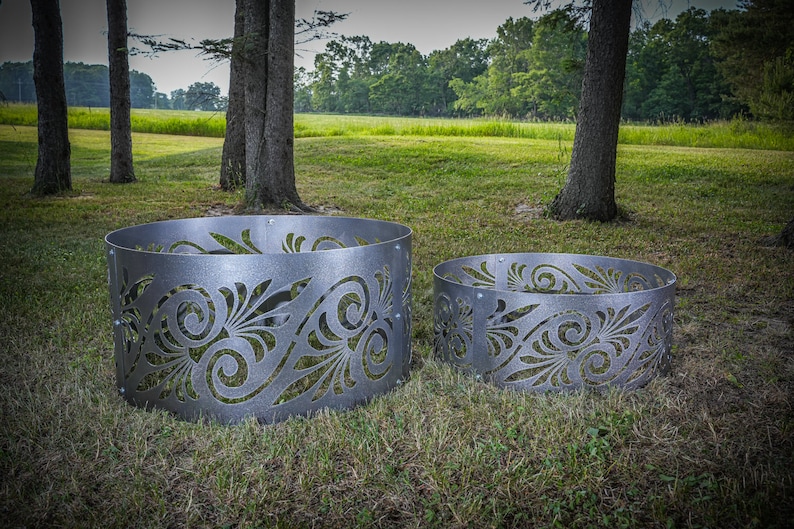 May include: Two circular metal fire pit rings with a silver-toned finish and a cut-out pattern featuring swirling, leaf-like designs. The rings are constructed from curved metal panels joined by visible rivets, creating a decorative barrier.