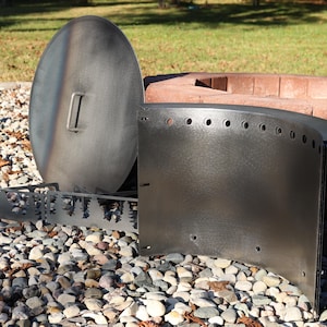 Close-up of a DIY Corten steel smokeless fire ring kit with pre-cut holes and welded lip, solid steel vent blocks, and solid lid with single handle in front of stacked retaining wall blocks on gravel patio.