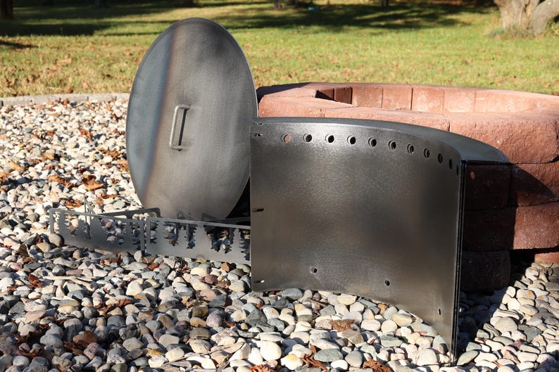 Close-up of a DIY Corten steel smokeless fire ring kit with pre-cut holes and welded lip, solid steel vent blocks, and solid lid with single handle in front of stacked retaining wall blocks on gravel patio.