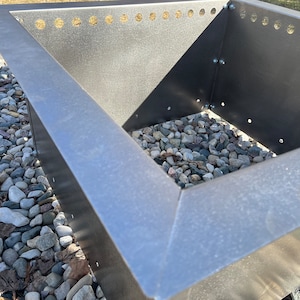 May include: Close-up of a square metal fire pit with a raised edge. The fire pit is filled with small gray rocks.