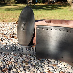 Close-up of a DIY smokeless fire ring in Corten steel kit with pre-cut holes and welded lip, solid steel vent blocks, and solid lid with single handle in front of stacked retaining wall blocks on gravel patio in a backyard with warm sun and shadow on the fire ring.