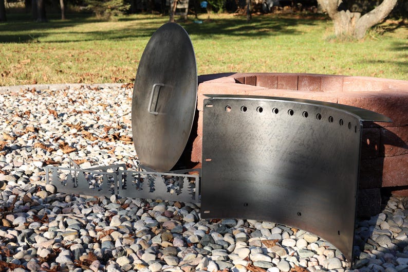 Close-up of a DIY smokeless fire ring in Corten steel kit with pre-cut holes and welded lip, solid steel vent blocks, and solid lid with single handle in front of stacked retaining wall blocks on gravel patio in a backyard with warm sun and shadow on the fire ring.