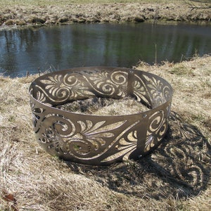 May include: A circular metal fire pit ring featuring a laser-cut pattern of swirling scrolls and leaf-like shapes. The structure is composed of multiple curved steel panels connected by visible bolts, creating a continuous decorative border.