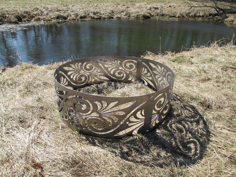 May include: A circular metal fire pit ring featuring a laser-cut pattern of swirling scrolls and leaf-like shapes. The structure is composed of multiple curved steel panels connected by visible bolts, creating a continuous decorative border.