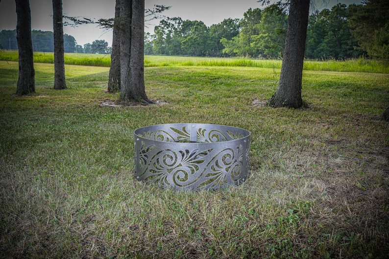May include: A circular metal fire pit ring featuring a laser-cut decorative pattern of swirling leaves and organic shapes. The silver-toned steel structure is composed of multiple panels connected by visible bolts, forming a continuous cylinder.