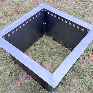 May include: A square metal fire pit with a black finish and holes around the edges. The fire pit is sitting on a grassy surface.