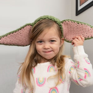 Op de afbeelding: Een jong meisje draagt ​​een roze en groene gehaakte hoed in de vorm van oren van een personage. De hoed heeft een groene bovenkant en roze oren. Het meisje draagt ​​een witte trui met een regenboogpatroon.
