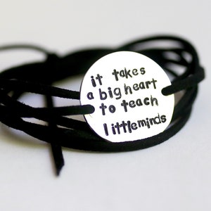 Op de afbeelding: Een zwart leren armband met een zilveren bedel waarop staat "het kost een groot hart om kleine geesten te onderwijzen".