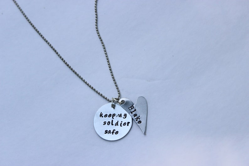 May include: Silver necklace with two charms. One charm is a round disc with the text "keep my soldier safe". The other charm is a heart-shaped disc with the name "Blake" inscribed on it.