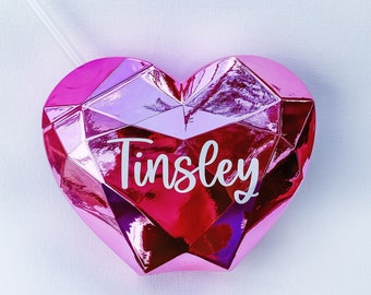 May include: Pink heart-shaped tumbler with a white straw and the name "Tinsley" printed on the front.