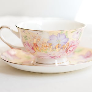 May include: A floral teacup and saucer set with a light yellow background and pink and purple flowers. The teacup has the name "Karen" in gold script. The set has gold trim.