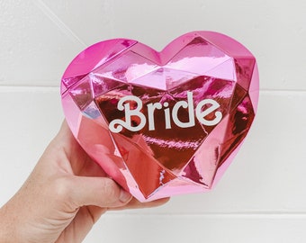 May include: Pink heart-shaped box with a shiny, metallic finish and the word "Bride" in white lettering.