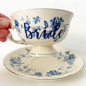 May include: Cream-colored teacup and saucer set with blue floral patterns and the word "Bride" in blue script. The teacup has a decorative handle and sits on a matching saucer with a silver rim. A classic and elegant design.