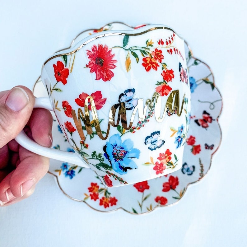 May include: A white teacup and saucer with a floral pattern and gold trim. The teacup has the word "Mama" written in gold lettering.