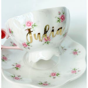 May include: White teacup and saucer set with a floral rose pattern in pink and green. The teacup has the name "Julia" in gold script. The handle is pink.